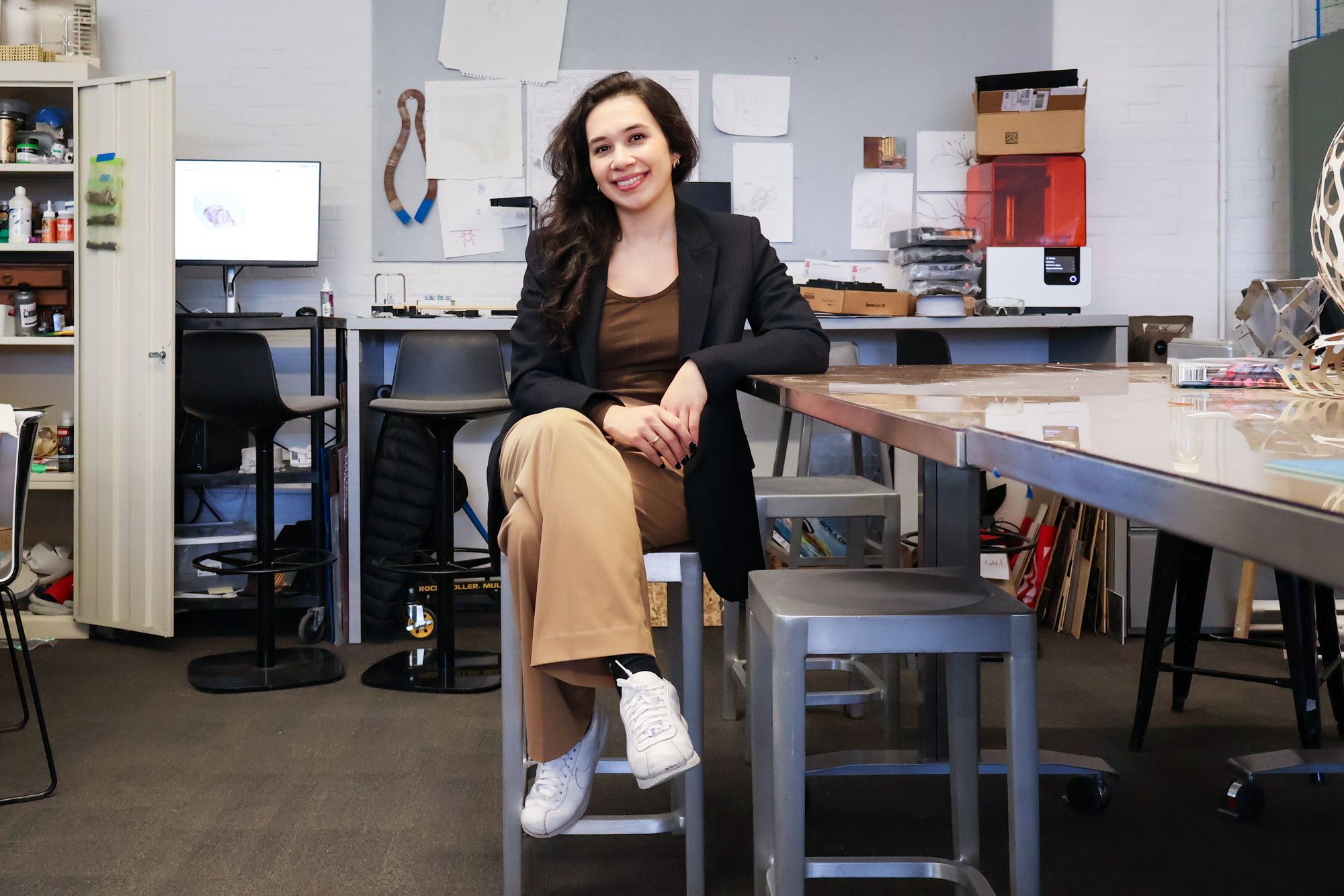 Young woman sitting on a stool in a studio or makerspace, smiling at the camera. She is wearing tan pants, a brown top, a black blazer, and white sneakers. Behind her are worktables, chairs, shelves with supplies, and sketches pinned to the wall.